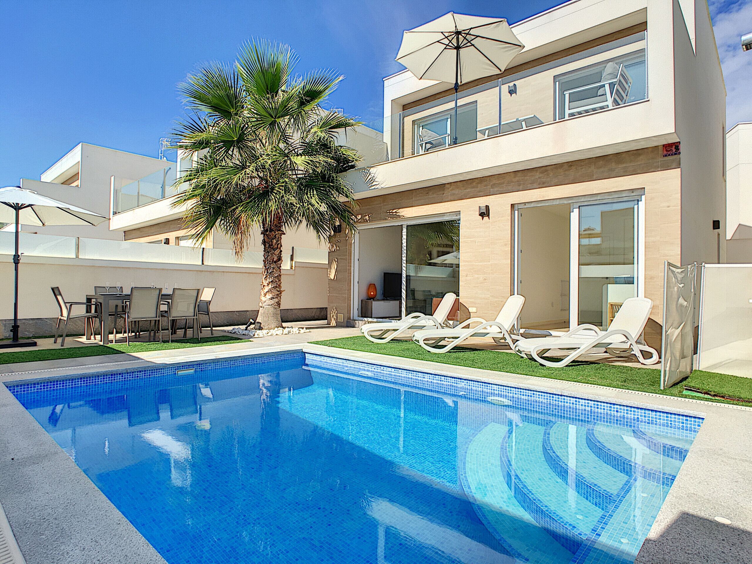 Un área de piscina moderna con una piscina azul cristalina, tumbonas, una palmera y un edificio de dos pisos con ventanas amplias y un gran ventanal corredero.