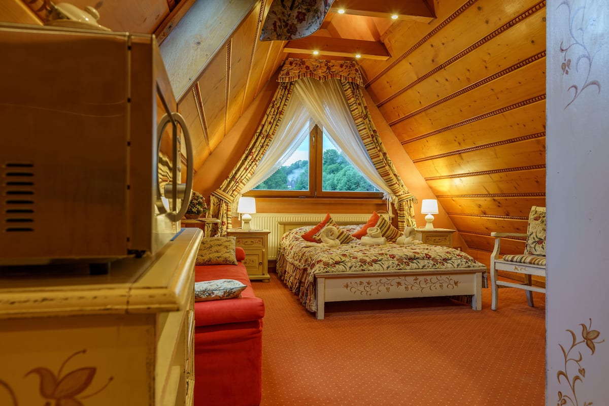 Une chambre avec un plafond en pente, un lit avec une literie à motifs floraux, une chaise rouge et une fenêtre avec des rideaux.