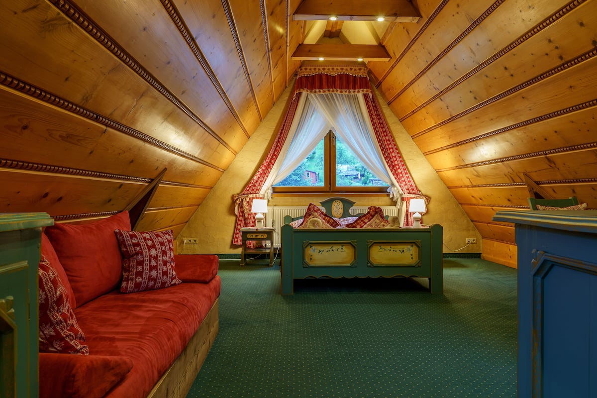 Une chambre mansardée chaleureuse avec un plafond en pente en bois, un lit à baldaquin, un canapé rouge et une petite table avec une lampe.