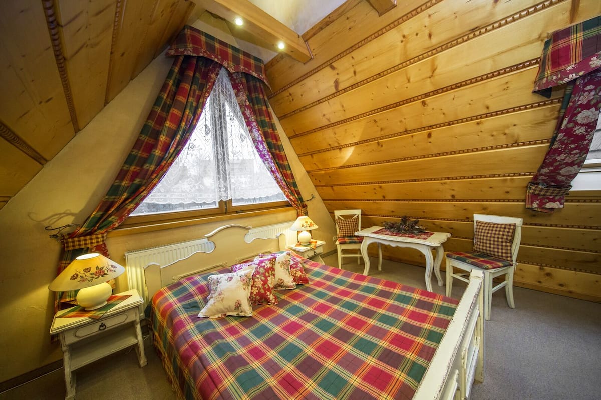 Une chambre mansardée chaleureuse, avec un lit, deux chaises, une petite table et une fenêtre avec des rideaux.