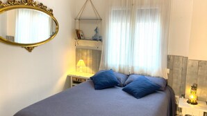 Une chambre avec un lit, une table de chevet avec une lampe, une étagère murale avec des livres et un ours en peluche, et un miroir au mur.
