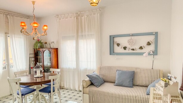 Un salon avec un canapé à rayures, une table basse en verre, une table à manger avec des chaises, un lustre et un cadre photo fixé au mur.