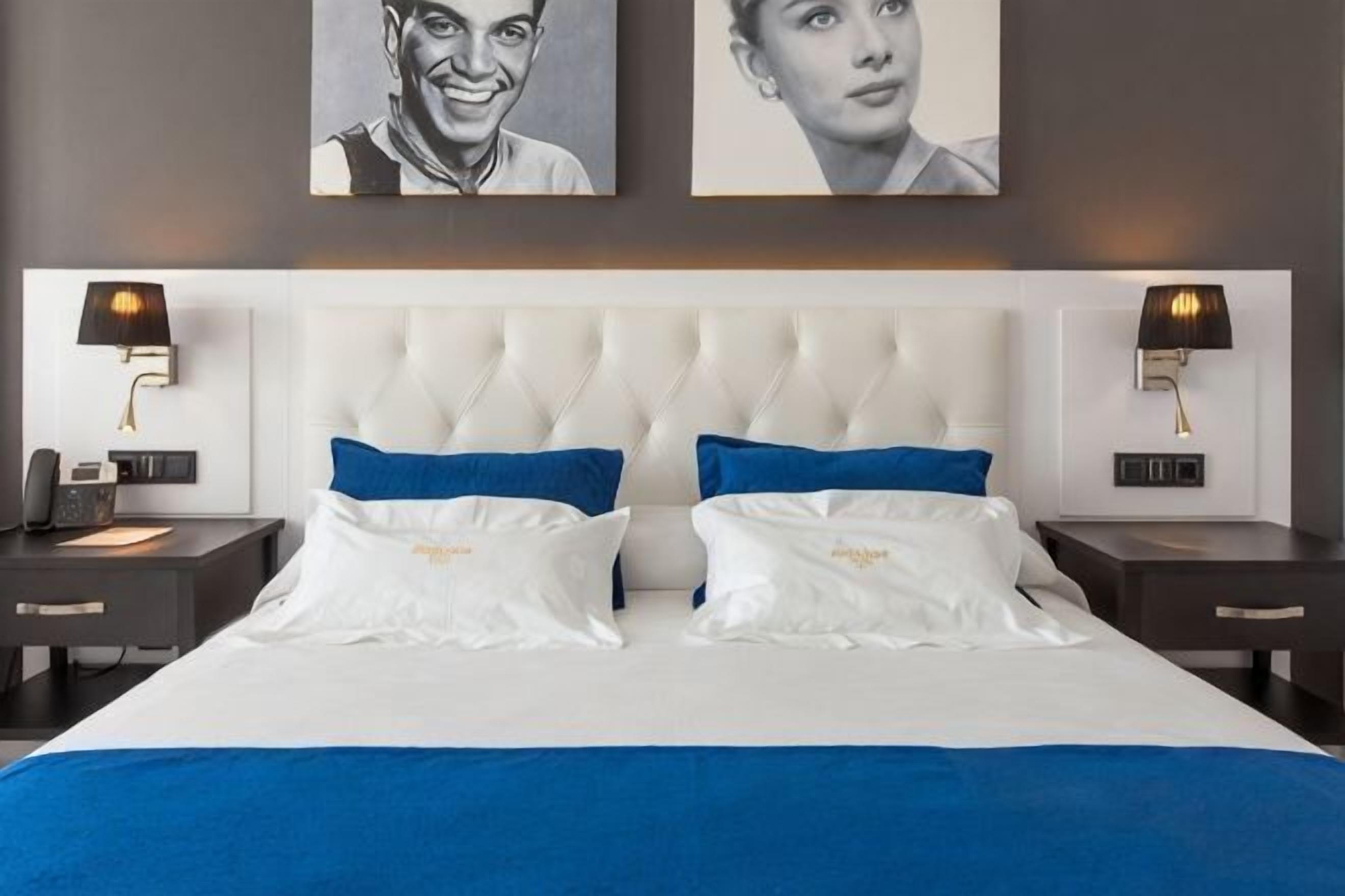 A hotel room with a large bed, two bedside tables, a nightstand, a wall-mounted lamp, and two framed pictures on the wall.