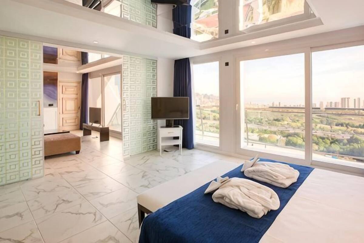 A hotel room with a bed, a TV, a balcony with a city view, and a large window.