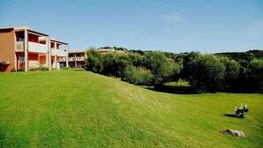 A building with multiple floors and balconies, surrounded by a well-maintained grassy area and dense shrubbery. - Hotel Club Baja Bianca (San Teodoro)