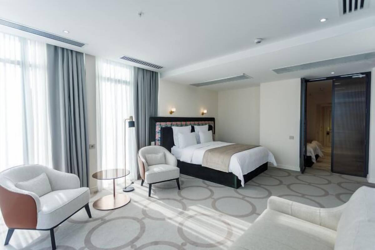 A modern hotel room with a large bed, two armchairs, a small round table, and a view of the hallway.