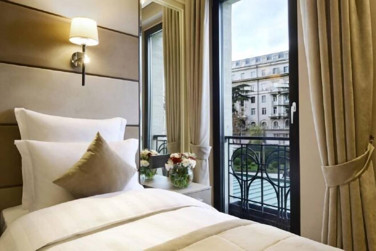 A hotel room with a bed, a nightstand, a vase with flowers, and a view of a balcony with a railing.