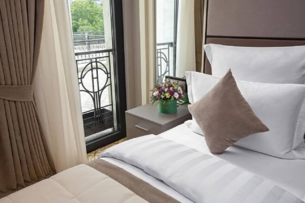 A hotel room with a bed, a nightstand, a window with a view of a balcony, and a vase with flowers.