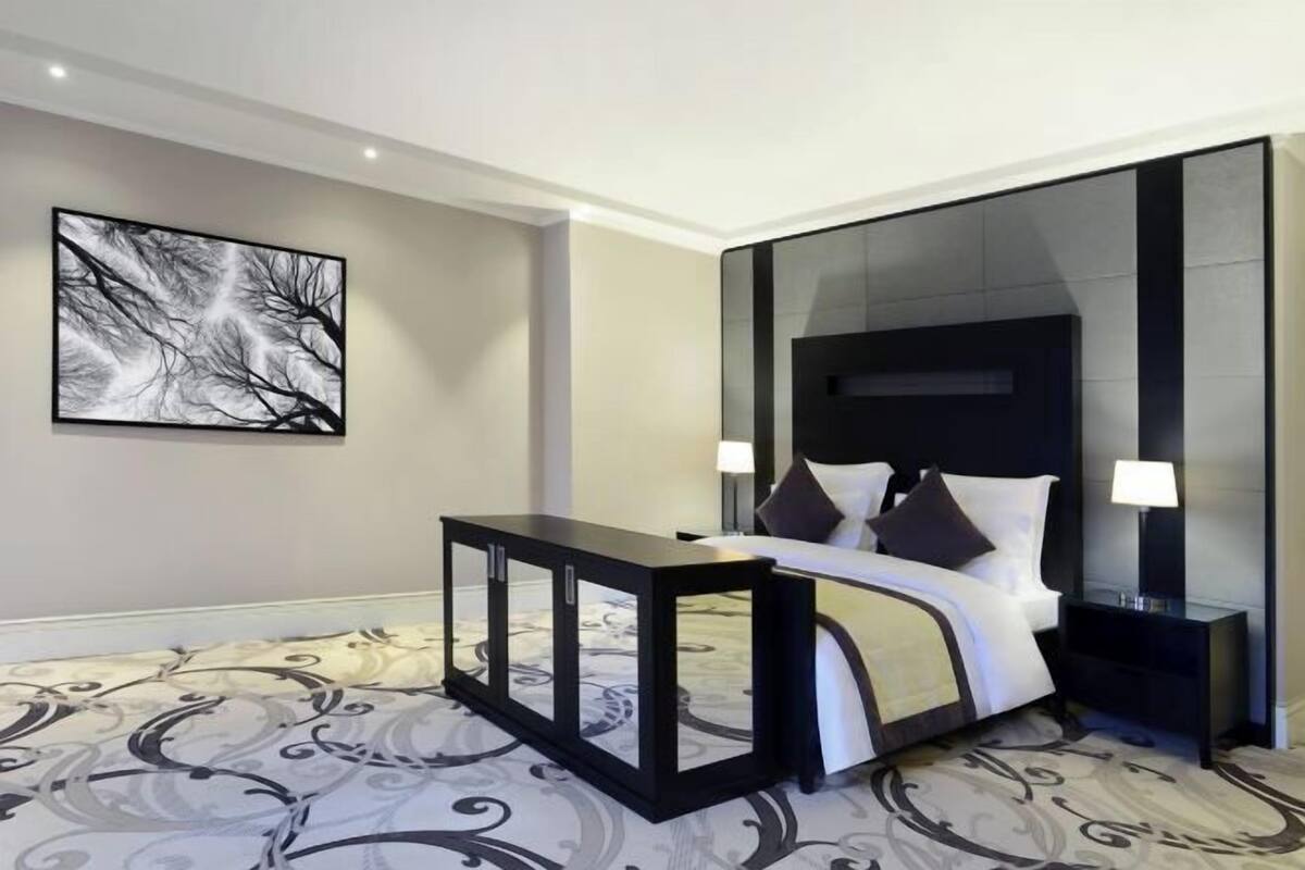 A modern hotel room with a bed, bedside tables, a large abstract painting, and a patterned carpet.