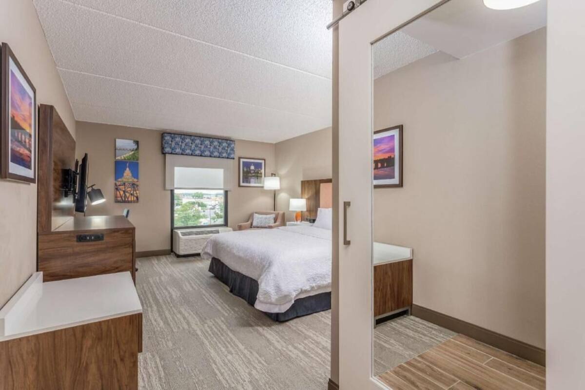 A hotel room with a bed, bedside table, dresser, window, and artwork on the walls.