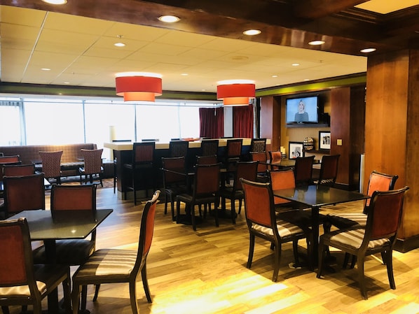 A hotel dining area with multiple tables and chairs, a bar, and a television screen. - Hampton Inn Harrisburg-east (Harrisburg)