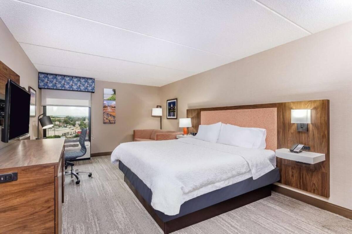 A hotel room with a bed, desk, chair, and a view of the city.