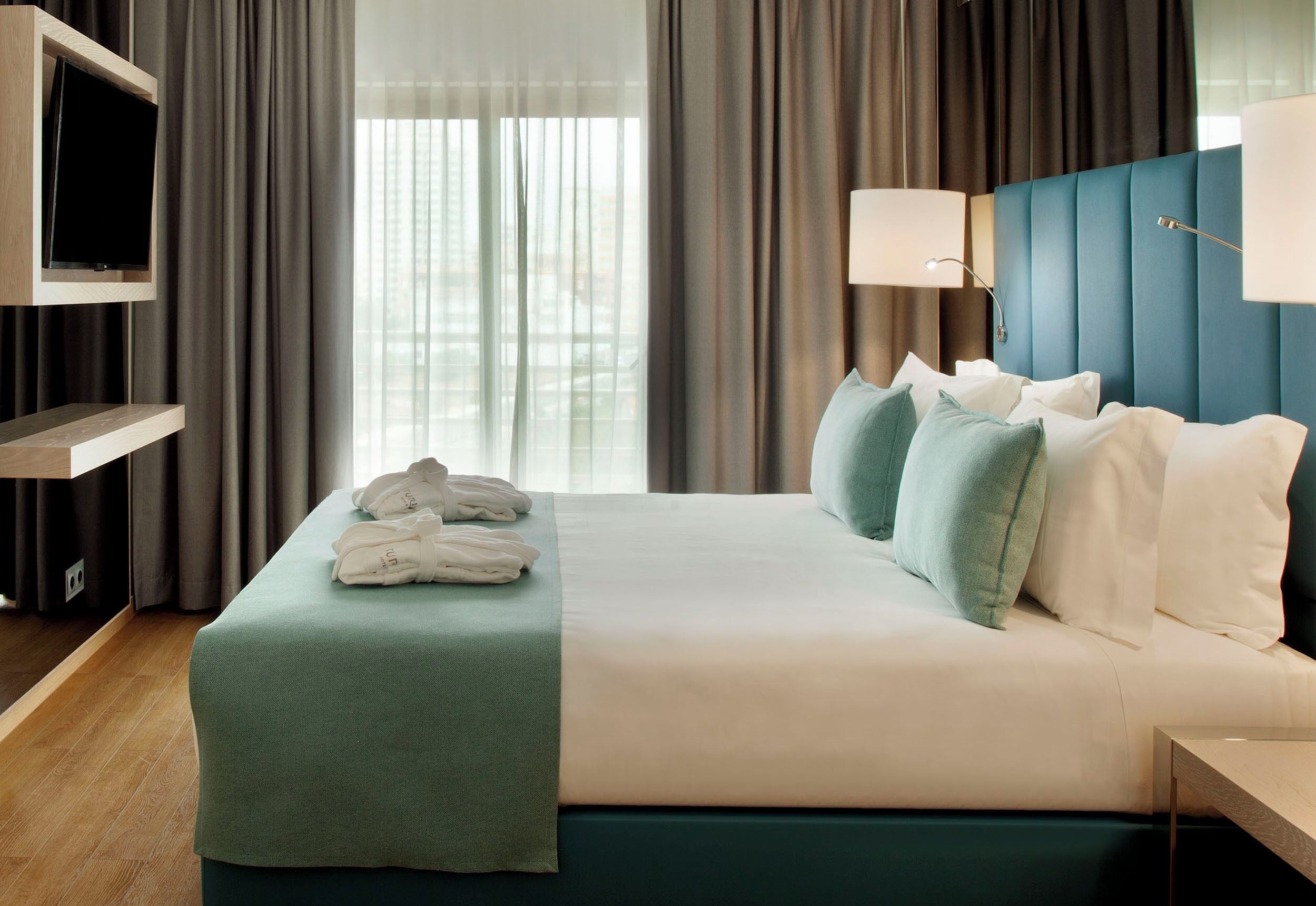 Habitación de hotel con cama, televisión montada en la pared, mesita de noche y ventana con cortinas.