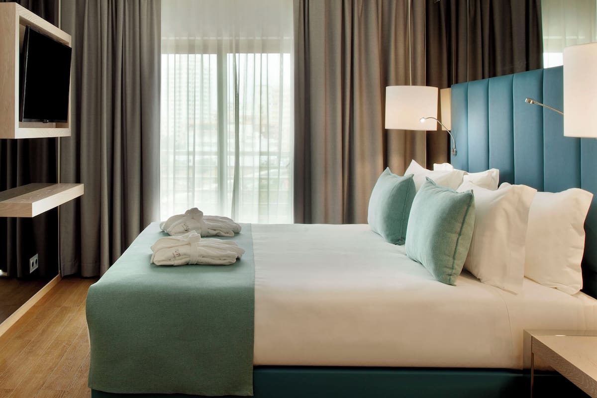 Quarto de hotel com cama, televisão na parede, mesa de cabeceira e janela com cortinas.