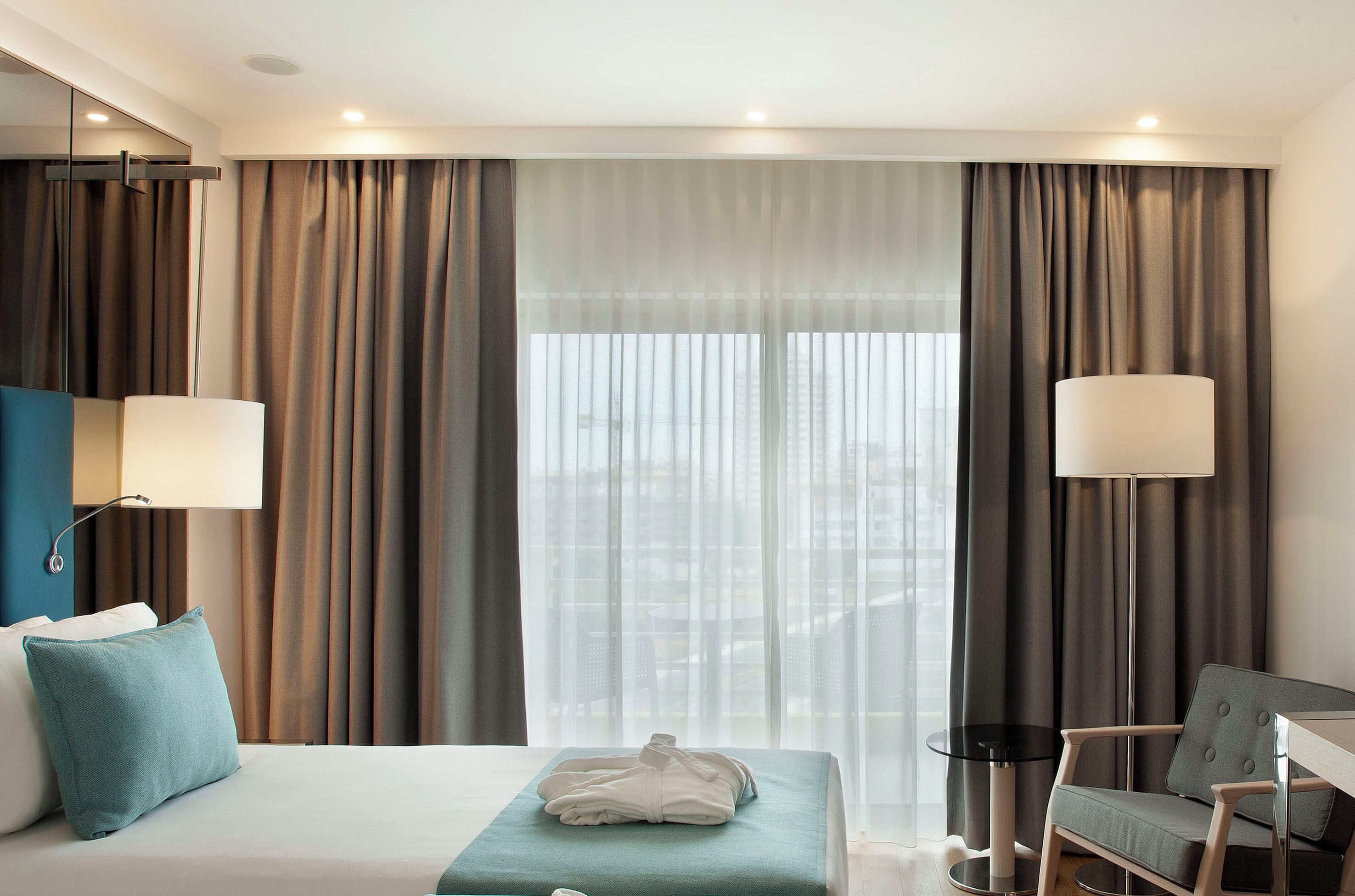 Habitación de hotel con cama, escritorio, silla y vista de la ciudad a través de cortinas traslúcidas.
