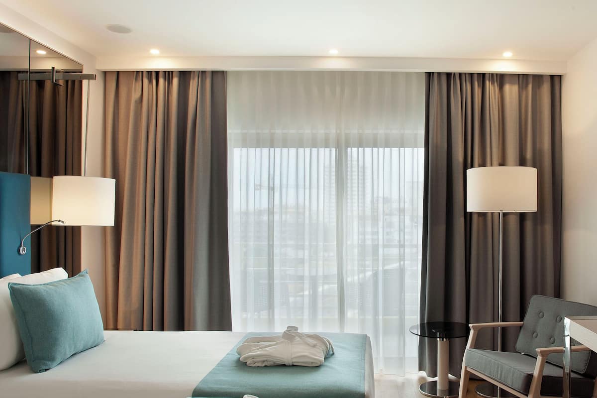 Quarto de hotel com cama, escrivaninha, cadeira e uma janela grande com cortinas.