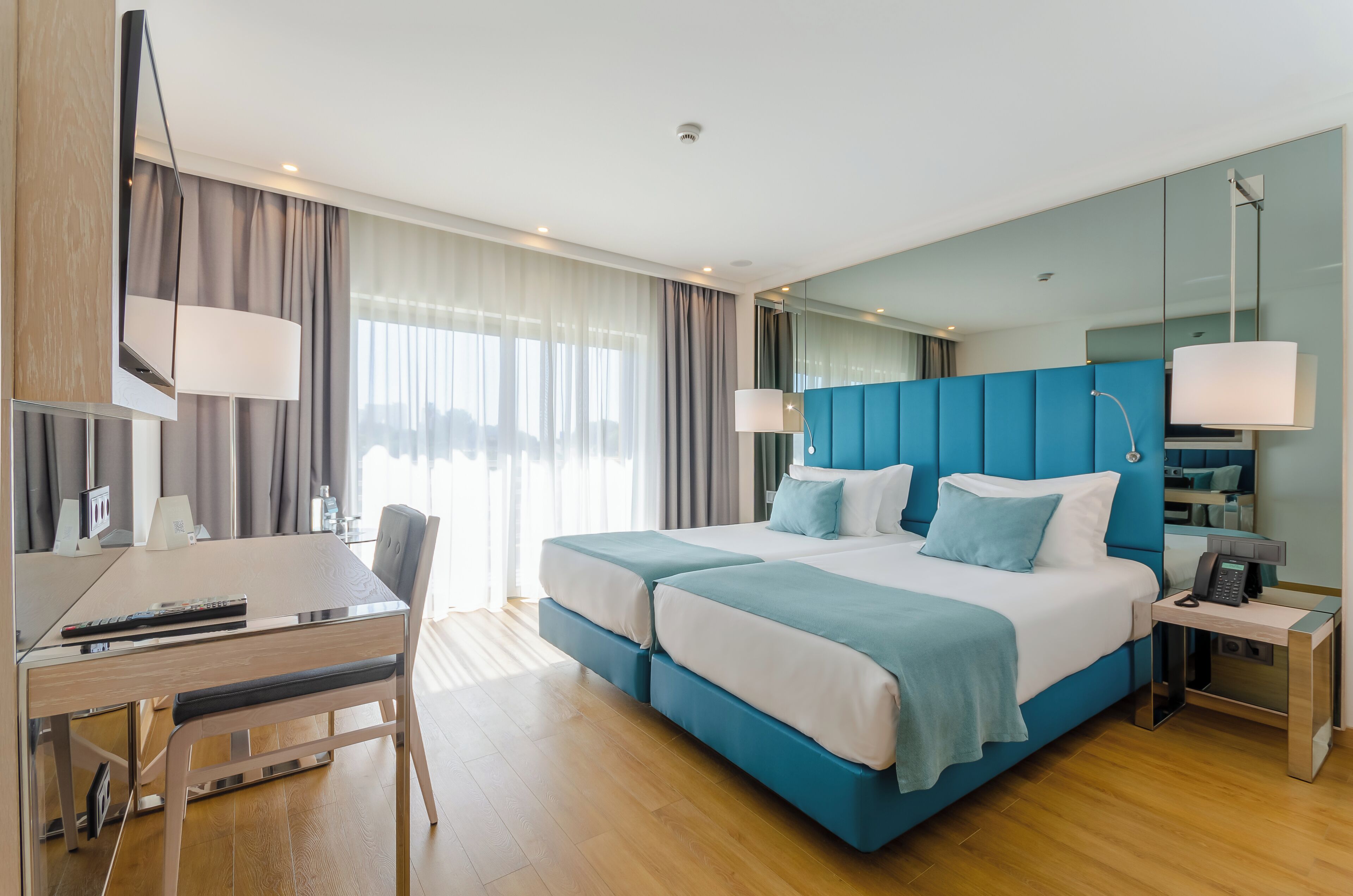 Habitación de hotel con cama, mesita de noche, lámpara, escritorio, silla y un gran ventanal con cortinas.