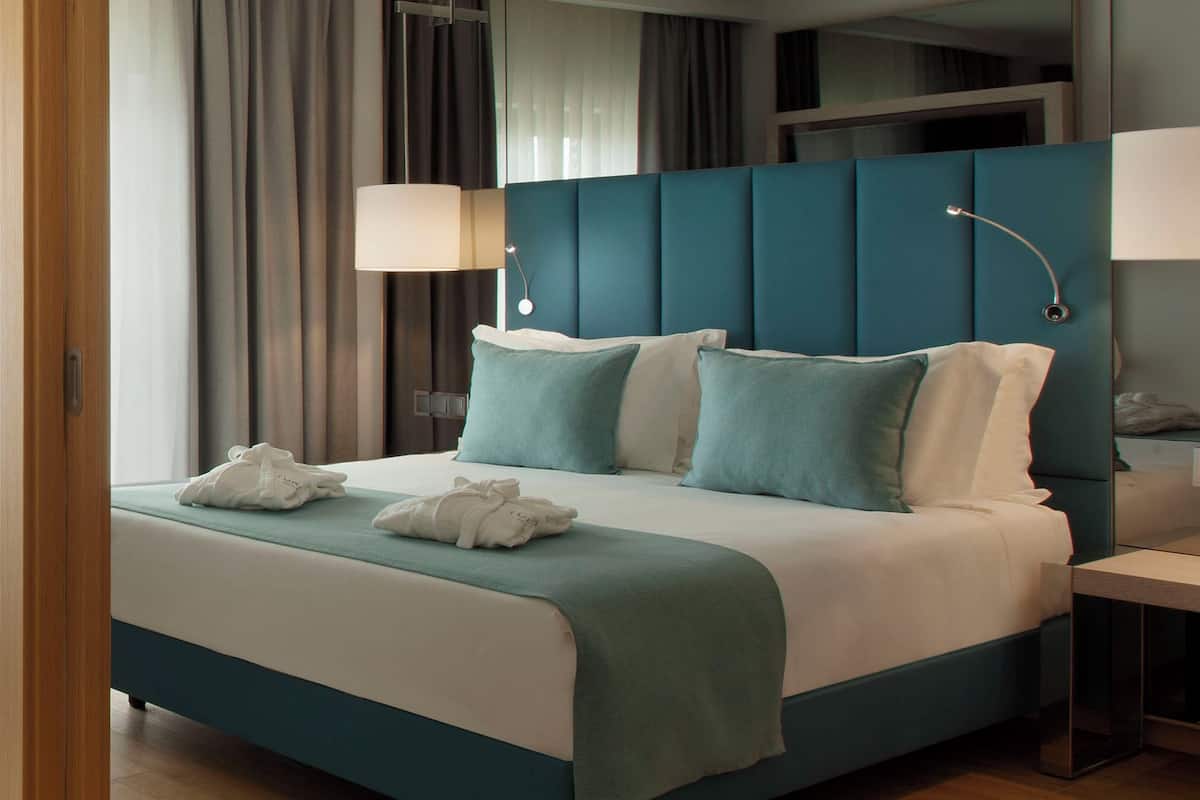 Quarto de hotel moderno com cabeceira estofada em tom azul-esverdeado, cama bem arrumada com lençóis brancos e mesa de cabeceira com abajur.