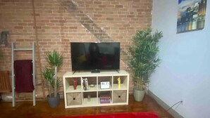 Una habitación con pared de ladrillo, un televisor de pantalla plana sobre un mueble blanco, una alfombra roja y una pintura de paisaje urbano en la pared.