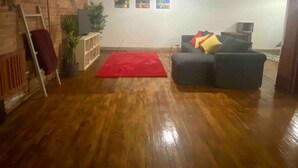 Un salón amplio con piso de madera, un tapete rojo, un sofá gris con cojines coloridos, un televisor y una pared de ladrillo.