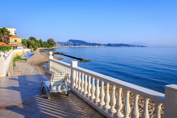 Un paseo marítimo con barandilla blanca, un banco y vistas al mar y colinas distantes.