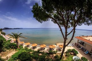 A beachside view with a clear sky, a large tree, and a row of beach umbrellas and lounge chairs. - Chryssi Akti Paradise (Zakynthos)