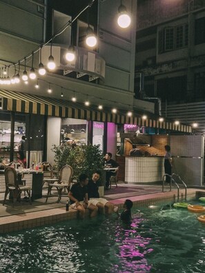 Zona de piscina interior con asientos y bar, rodeada de luces colgantes y una pared de ladrillo.