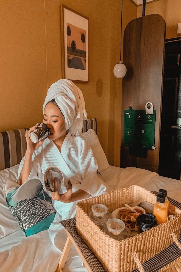A person sitting on a bed with a tray of food and drinks, a wooden door, and a wall-mounted coat rack. - Josh Hotel (Bangkok)