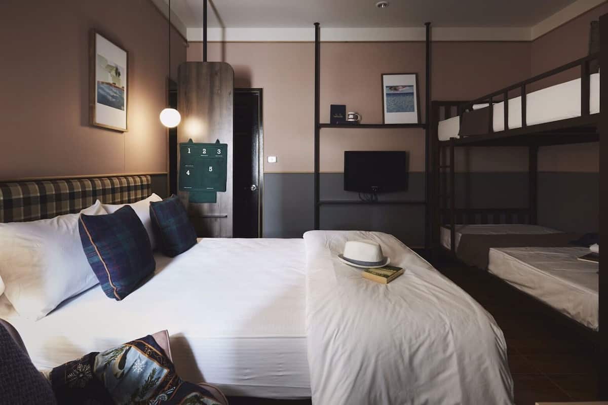 Ein Hotelzimmer mit einem großen Bett, einem kleineren Bett, Stockbetten, einem Schreibtisch mit Computer, einem Fernseher und einer wandmontierten Steuerung.