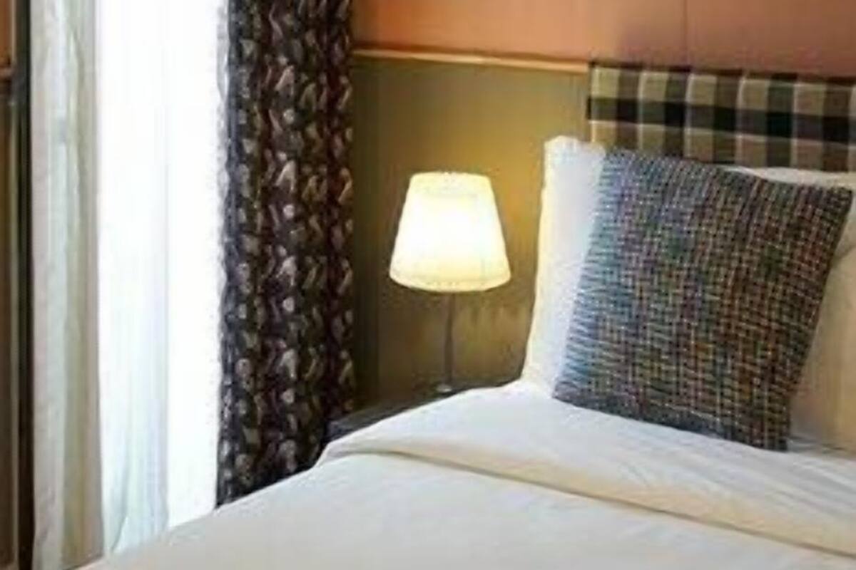 Ein Hotelzimmer mit einem Bett, einer Lampe, einem Fenster mit Vorhängen und einem Kopfteil.