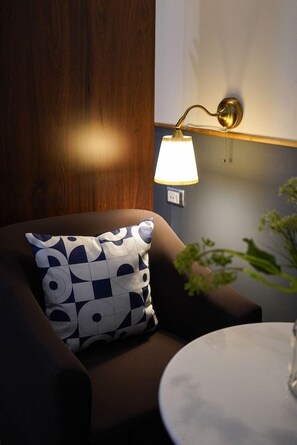 A room with a brown sofa, a patterned pillow, a wall-mounted lamp, and a round table with a vase. - Josh Hotel (Bangkok)
