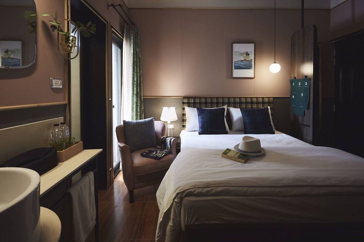 Ein Hotelzimmer mit Bett, Sessel, Nachttisch, Lampe, Spiegel, Fenster mit Vorhängen und einem an der Wand befestigten Regal mit Pflanze.