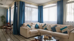 Un salon avec un canapé d’angle beige, des rideaux bleus et blancs, une table basse en verre et un tapis à motifs.