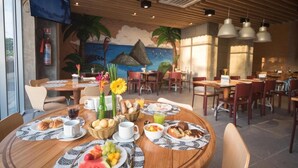 Ein Restaurant mit runden Tischen, Frühstücksangeboten und einem Wandgemälde einer Strandlandschaft.