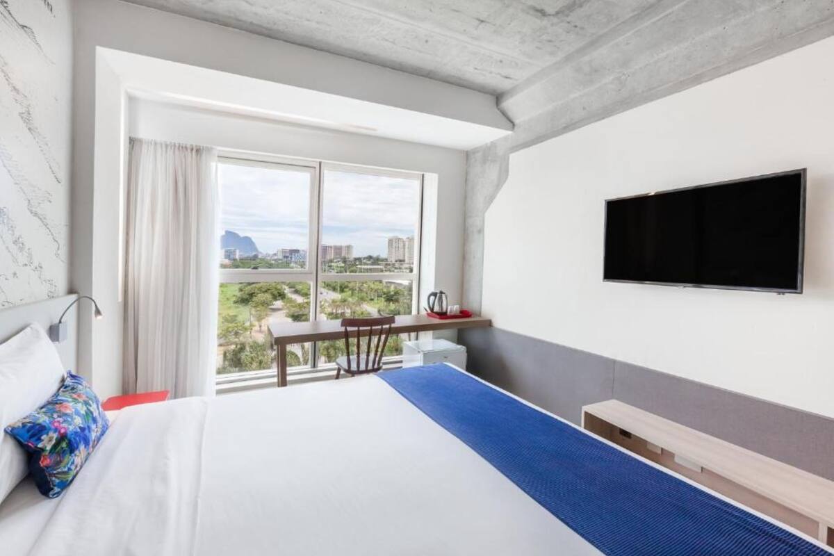 A hotel room with a bed, a desk, a chair, a TV, and a view of the city.