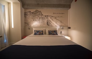 Une chambre d’hôtel avec un grand lit, deux coussins à motifs floraux, une fresque représentant un paysage urbain sur le mur et une valise rouge posée sur la table de chevet.