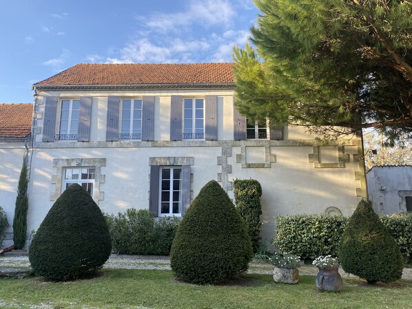 A two-story building with a tiled roof, multiple windows with shutters, and a well-maintained garden with trimmed hedges and a stone pathway.