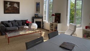 A spacious living room with a fireplace, a sofa, a dining table, and large windows offering a view of trees.