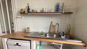 A compact kitchen with a dishwasher, a sink, and a wooden countertop. There are shelves above the sink holding various kitchen items.
