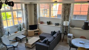 Un salón con sofá, televisión, mesa de centro y mesa de comedor con sillas. Hay ventanas con cortinas, una estantería con plantas y una lámpara.