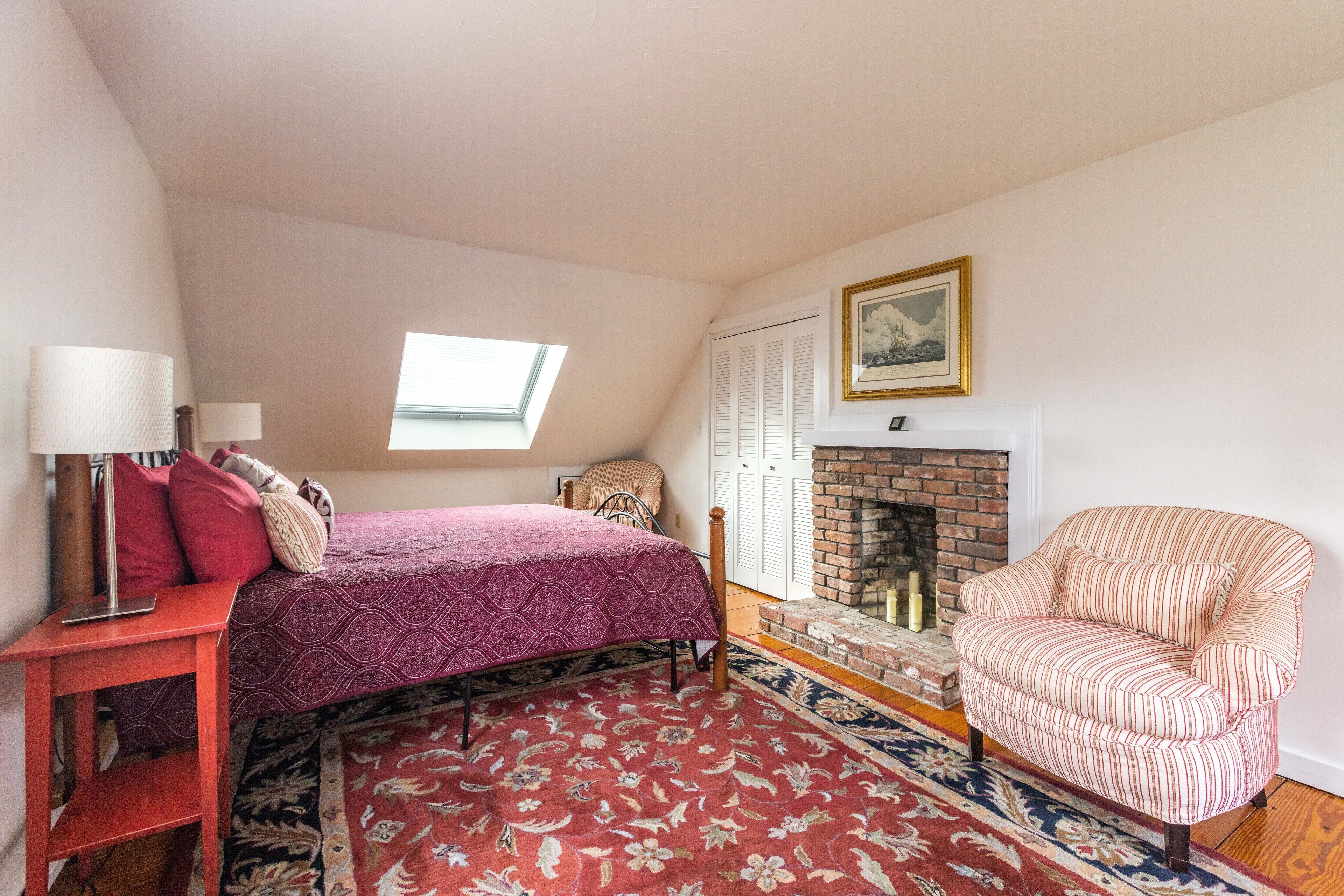 A bedroom with a bed, a chair, a fireplace, a window, and a door.
