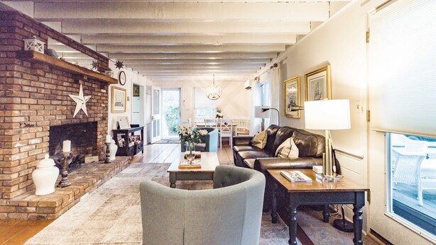 A spacious living room with a brick fireplace, a sofa, a coffee table, and a dining area in the background. - Charming Vineyard Saltbox + Ferry Tickets (Edgartown)