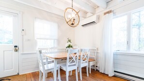A dining area with a wooden table and white chairs, a hanging light fixture, and a window with a view of trees. - Charming Vineyard Saltbox + Ferry Tickets (Edgartown)