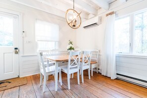 A dining area with a wooden table and white chairs, a hanging light fixture, and a window with a view of trees.