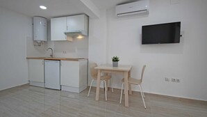 Una cucina compatta con mobili bianchi, lavastoviglie, lavello e cappa aspirante integrata. È presente un piccolo tavolo da pranzo con due sedie e una pianta in vaso. Un televisore a schermo piatto è montato a parete sopra la zona pranzo.