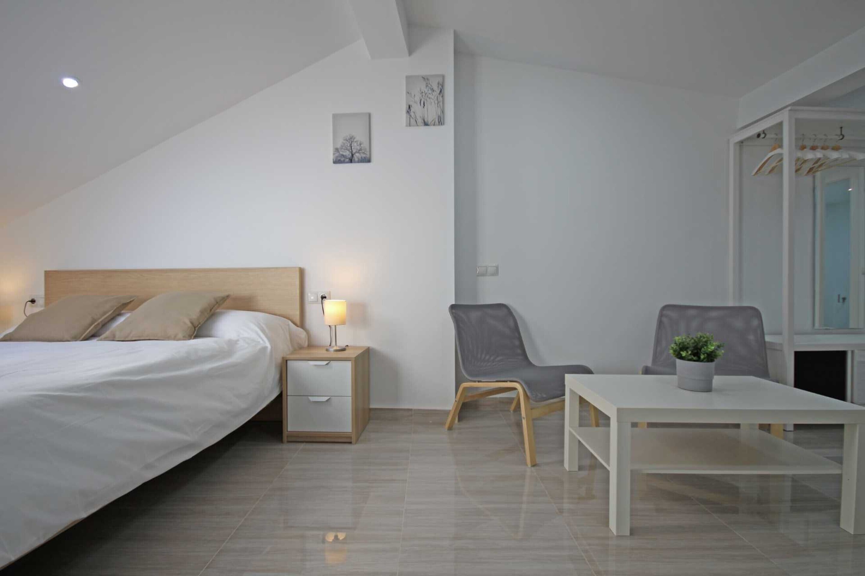 A modern bedroom with a bed, bedside table, lamp, two chairs, a small table, and a potted plant.