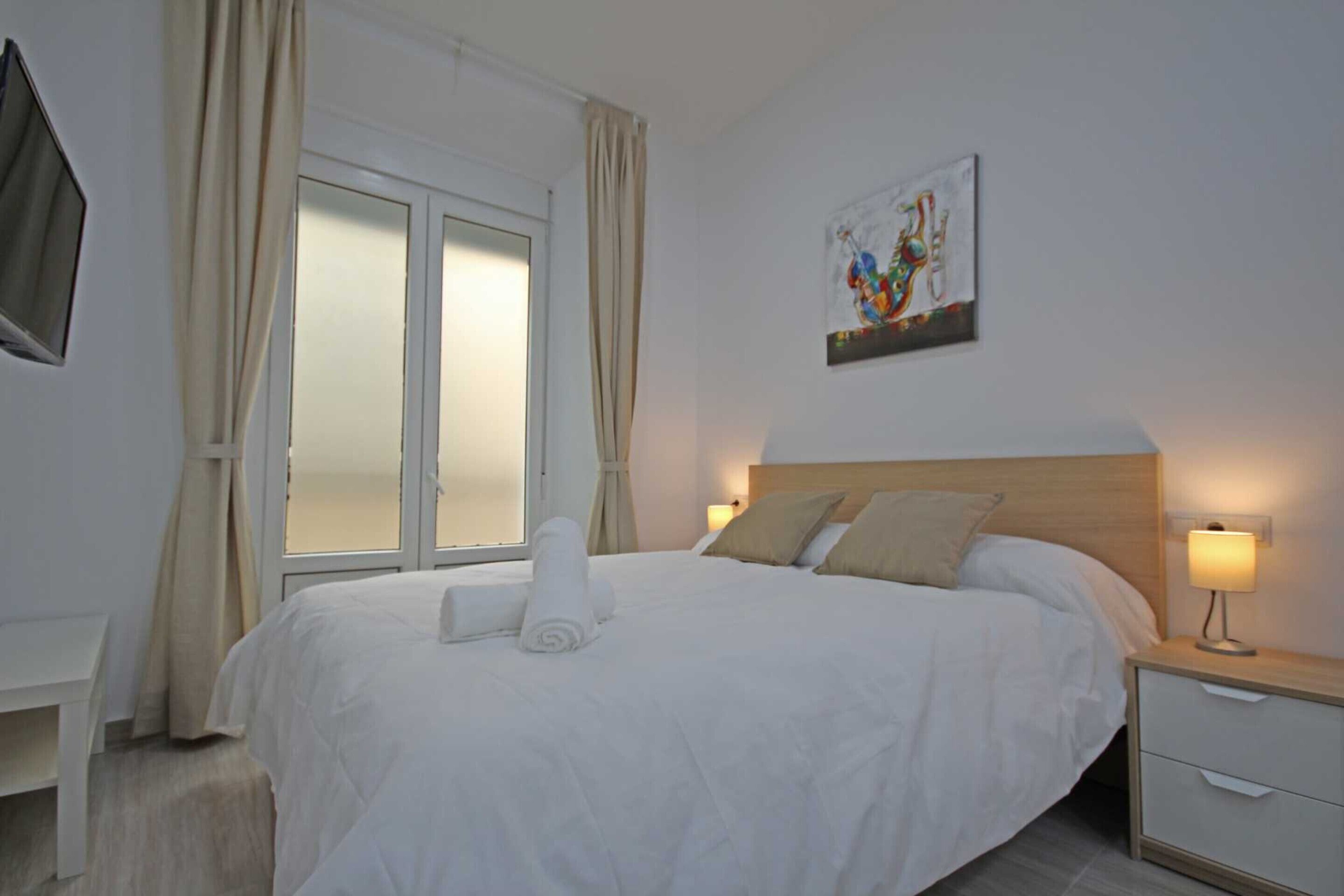 Una cama bien tendida con sábanas blancas y una toalla enrollada, una mesita de noche con una lámpara y una ventana con cortinas.