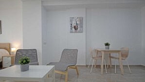 Una stanza moderna e minimalista con un letto bianco, una piccola scrivania e un tavolo da pranzo con sedie. Sulla scrivania c'è una pianta in vaso e al muro un quadro.