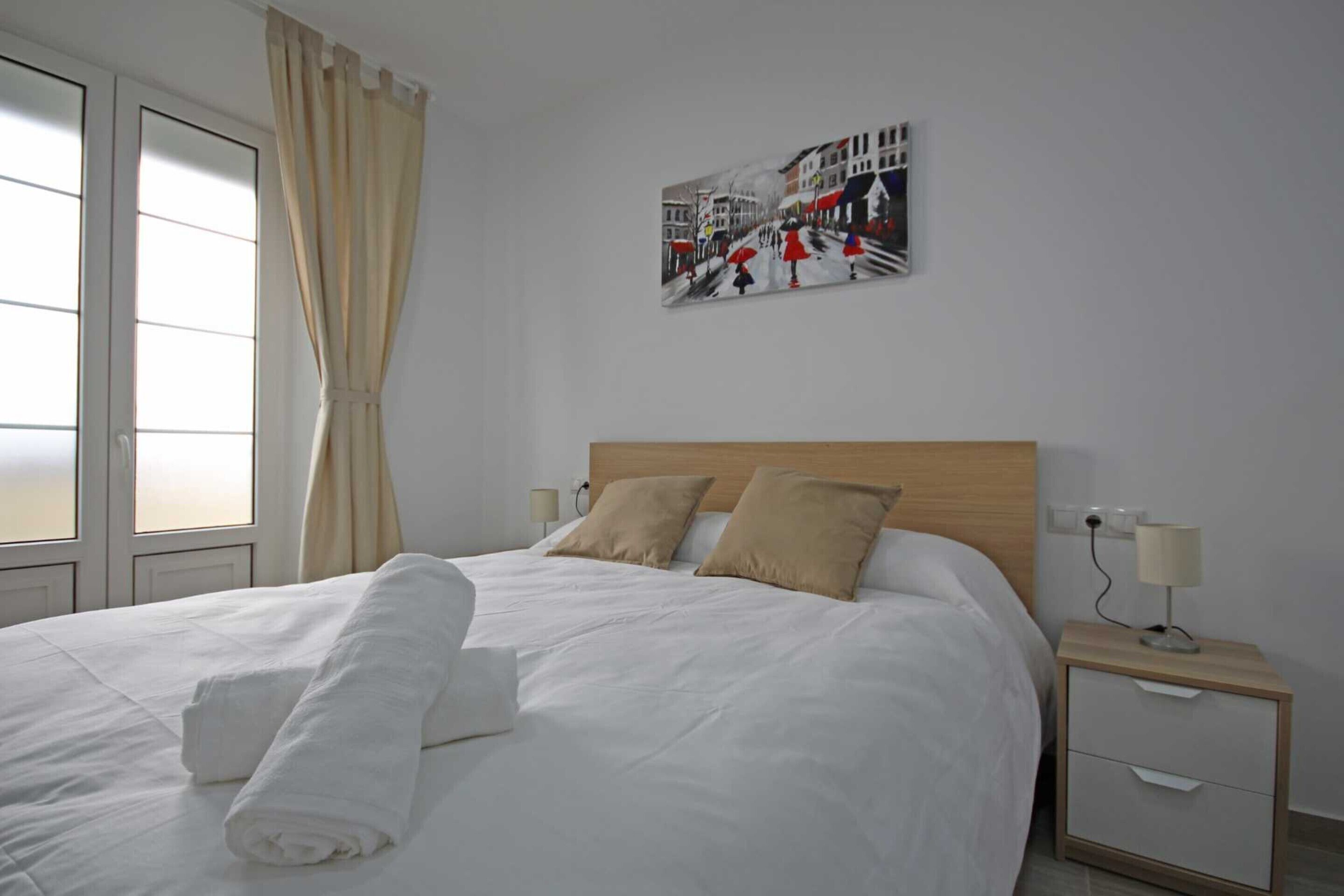 Ein ordentlich bezogenes Bett mit weißen Leinen und einem aufgerollten Handtuch, ein hölzernes Kopfteil, ein Nachttisch mit Lampe sowie ein Gemälde einer Straßenszene an der Wand.