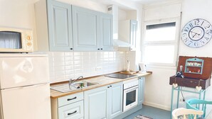 A kitchen with light blue cabinets, a white refrigerator, a sink, and a stove. There is a colorful rug on the floor and a clock on the wall.